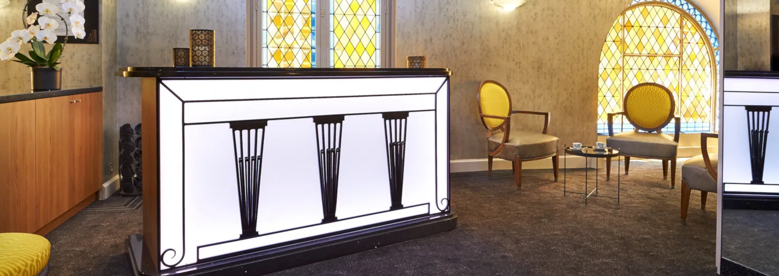 Modern hotel lobby with Art Deco design featuring a illuminated reception desk with black geometric patterns, yellow accent chairs, stained glass windows, and warm gold-toned wallpaper