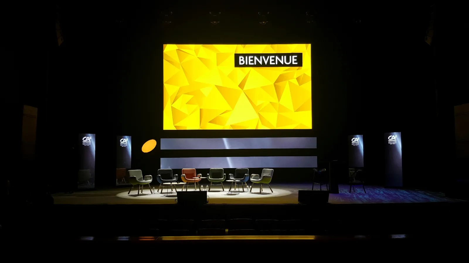 Conference stage with yellow welcome display showing 'BIENVENUE', six colorful chairs arranged in a semicircle, and branded panels with CA logos on either side