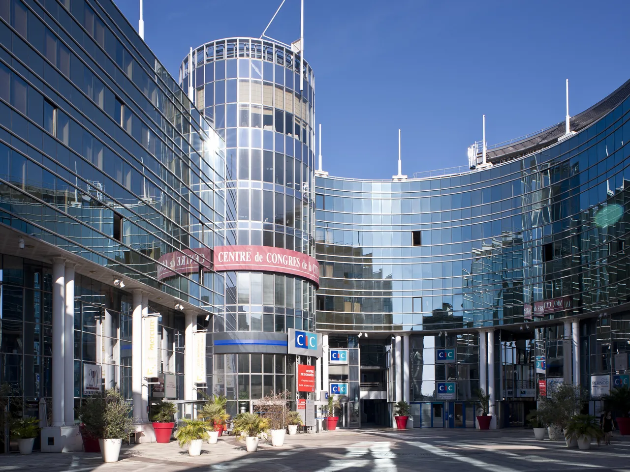 Modern architectural complex featuring a cylindrical glass tower with white framework, surrounded by curved blue-tinted buildings. The central plaza displays signage for 'Centre de Congrès' with decorative red planters and landscaping under clear blue sky