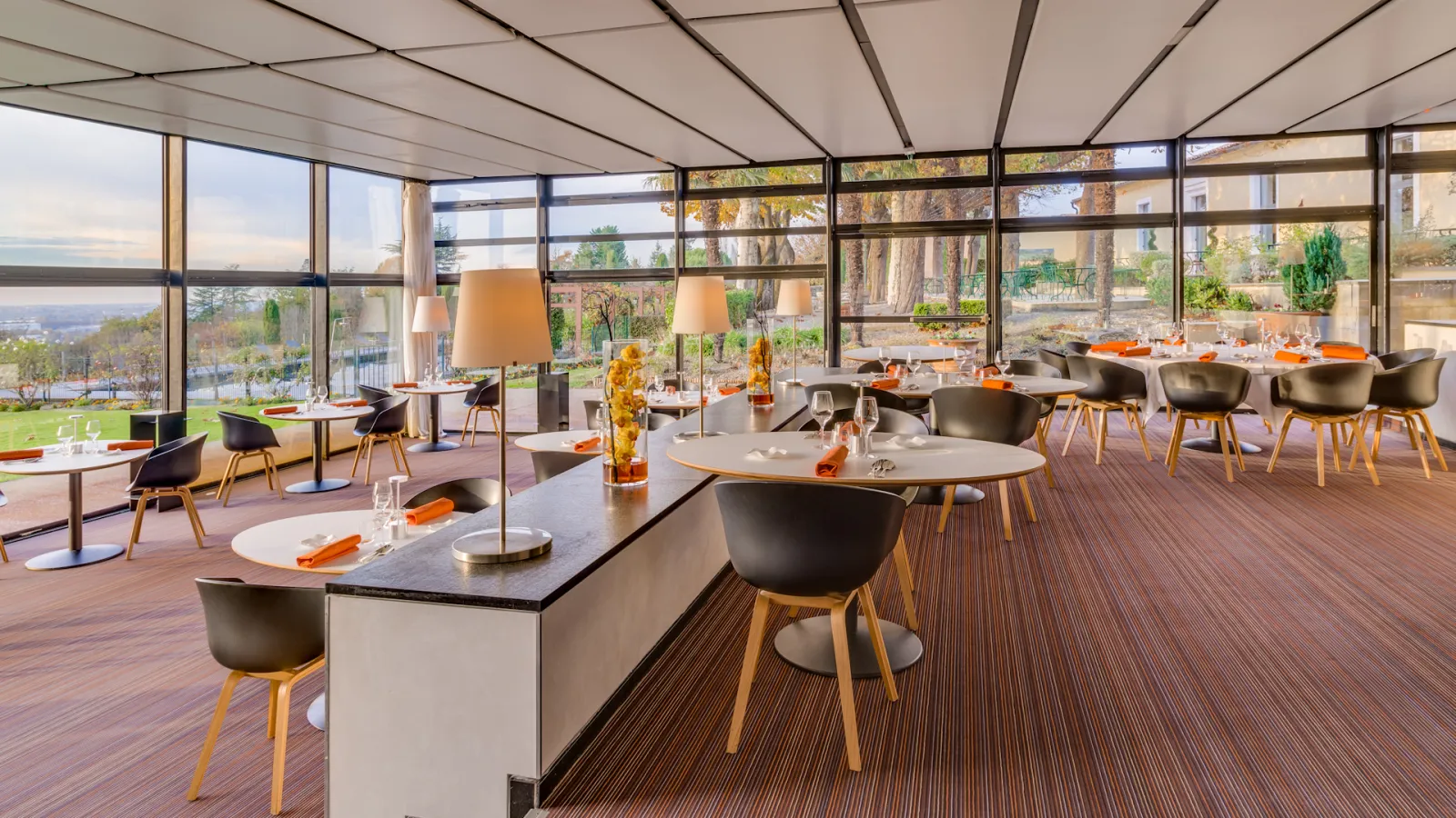 Modern restaurant dining room at Le Saint-James hotel in Bordeaux with floor-to-ceiling windows overlooking the landscape, featuring contemporary tables with black chairs and wooden legs, warm lighting, and elegant place settings with orange accents.