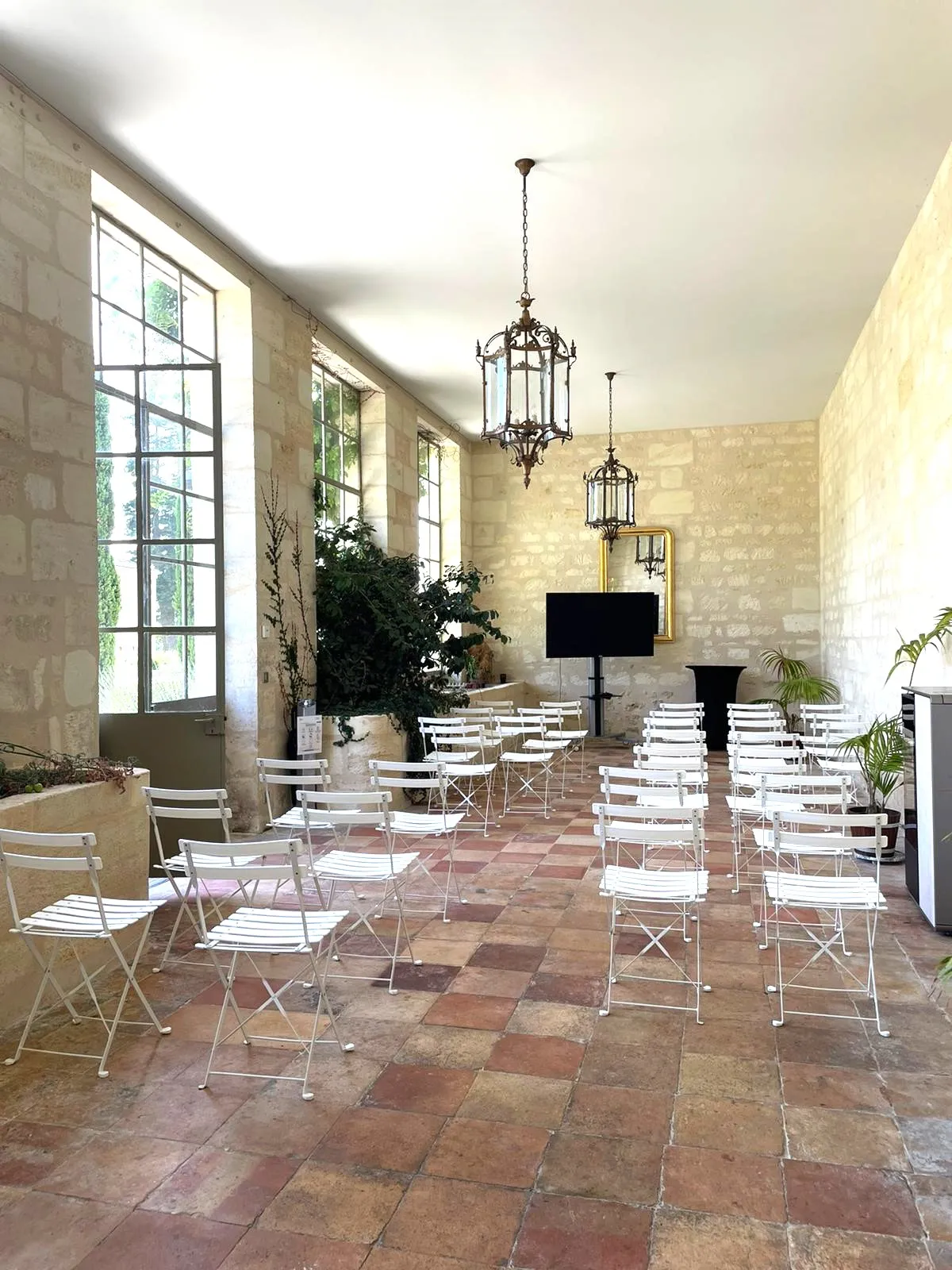 Spacious reception hall at Chateau Gassies in Bordeaux with terracotta tile flooring, white folding chairs arranged in rows, decorative hanging lanterns, large windows with gridded panes, exposed stone walls, potted plants, and a display screen.