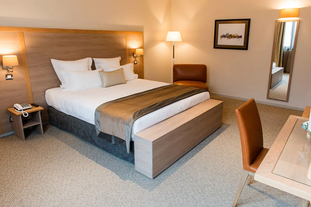 Modern hotel bedroom at Vatel Bordeaux featuring a king-size bed with white linens, wooden headboard with integrated lighting, bench at the foot of the bed, leather armchair, desk area, and ensuite bathroom visible through an open door.
