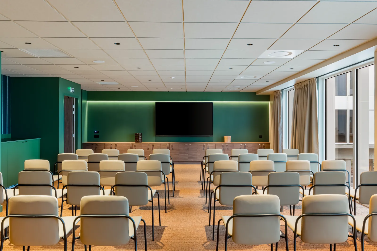 Modern conference room at Hampton By Hilton Bordeaux Mérignac with rows of gray upholstered chairs facing a large black screen mounted on a green accent wall with ambient lighting, wooden side cabinet, and floor-to-ceiling windows with beige curtains.