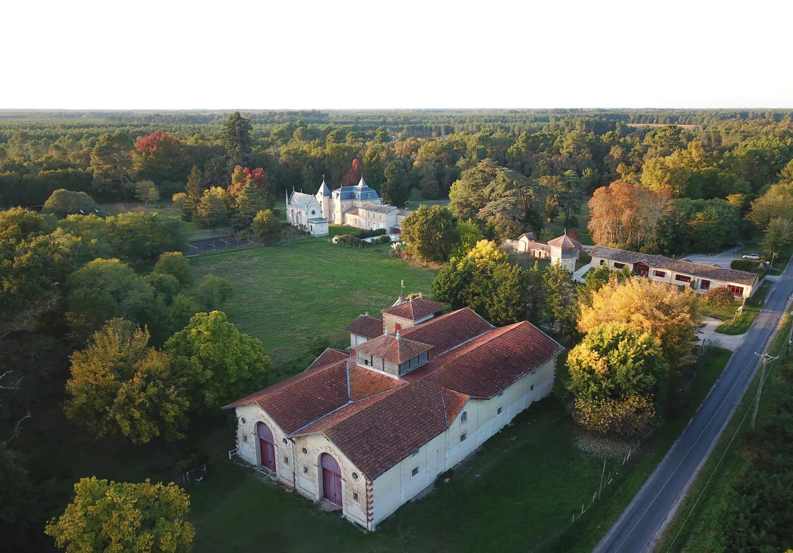 Aerial view of Château Léognan estate in Bordeaux, France, featuring a white fortified building with red-tiled roof in the foreground, a blue-roofed château in the background, surrounded by autumn-colored forests and green fields.