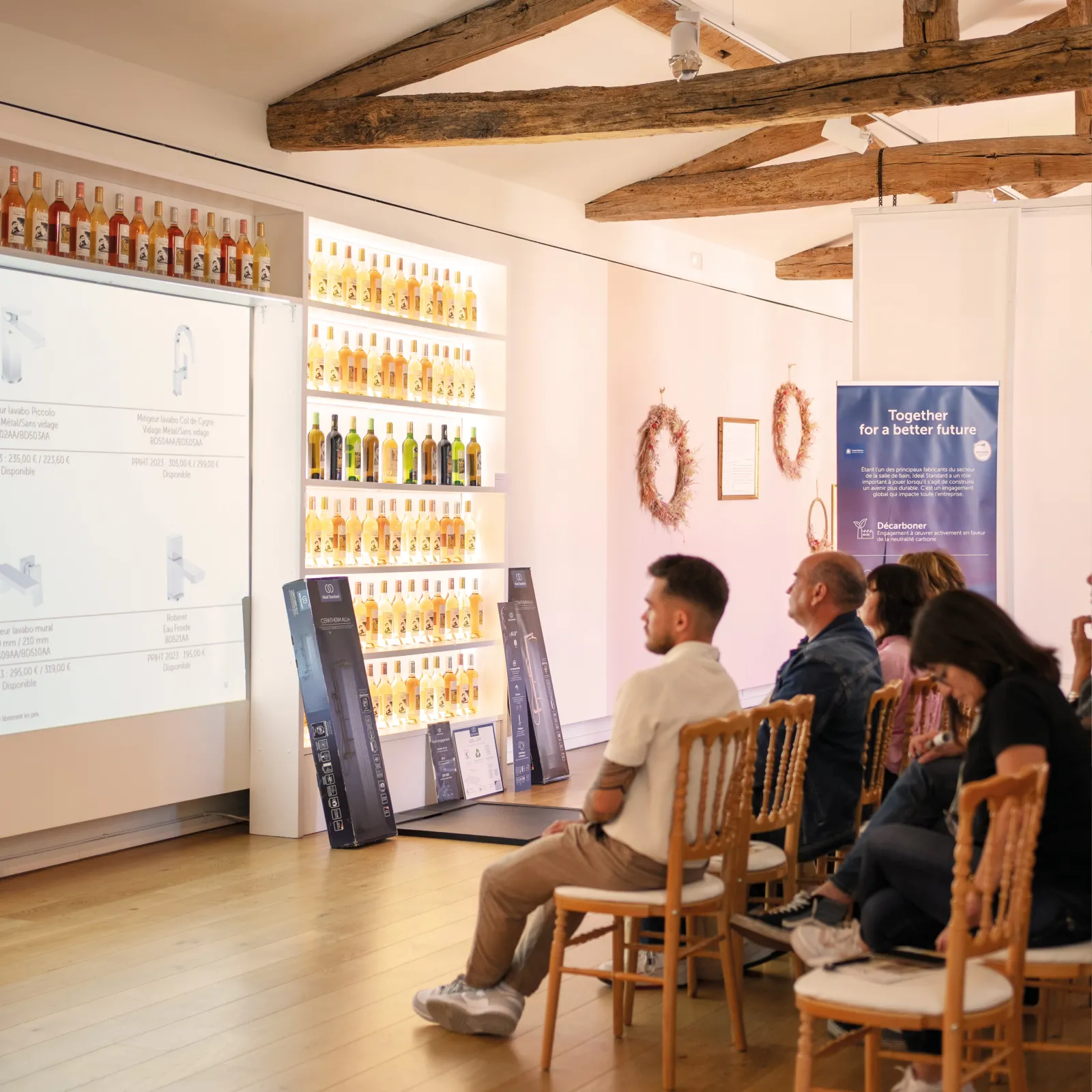 Visitors seated in a modern tasting room at Château Toulouse-Lautrec in Bordeaux, with exposed wooden beams, wine bottles displayed on illuminated shelves, and a presentation screen showing wine information.
