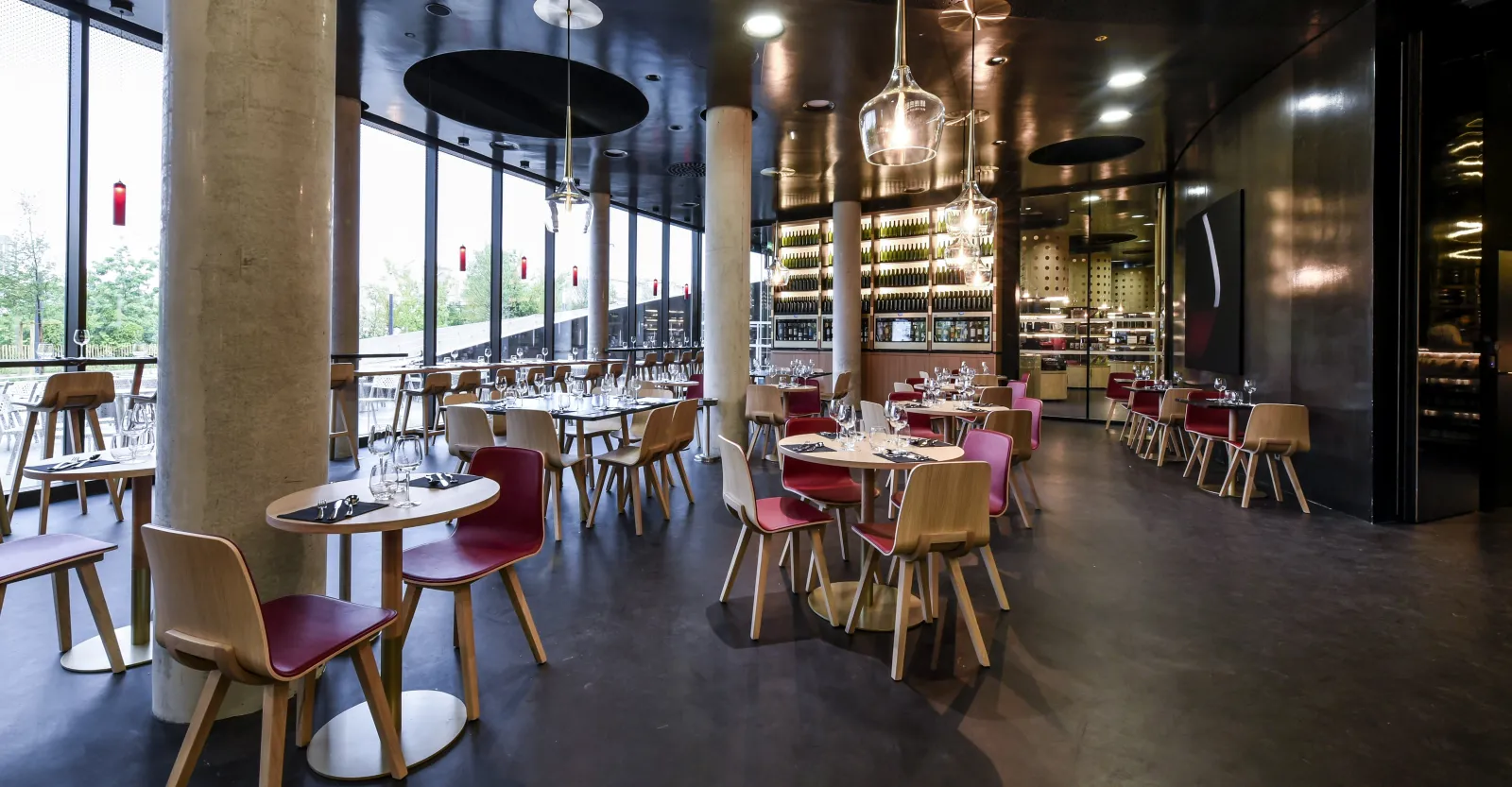 Modern restaurant interior with wooden chairs and burgundy seats arranged around small tables, featuring a sophisticated bar with extensive bottle selection, pendant lighting, and floor-to-ceiling windows overlooking greenery.