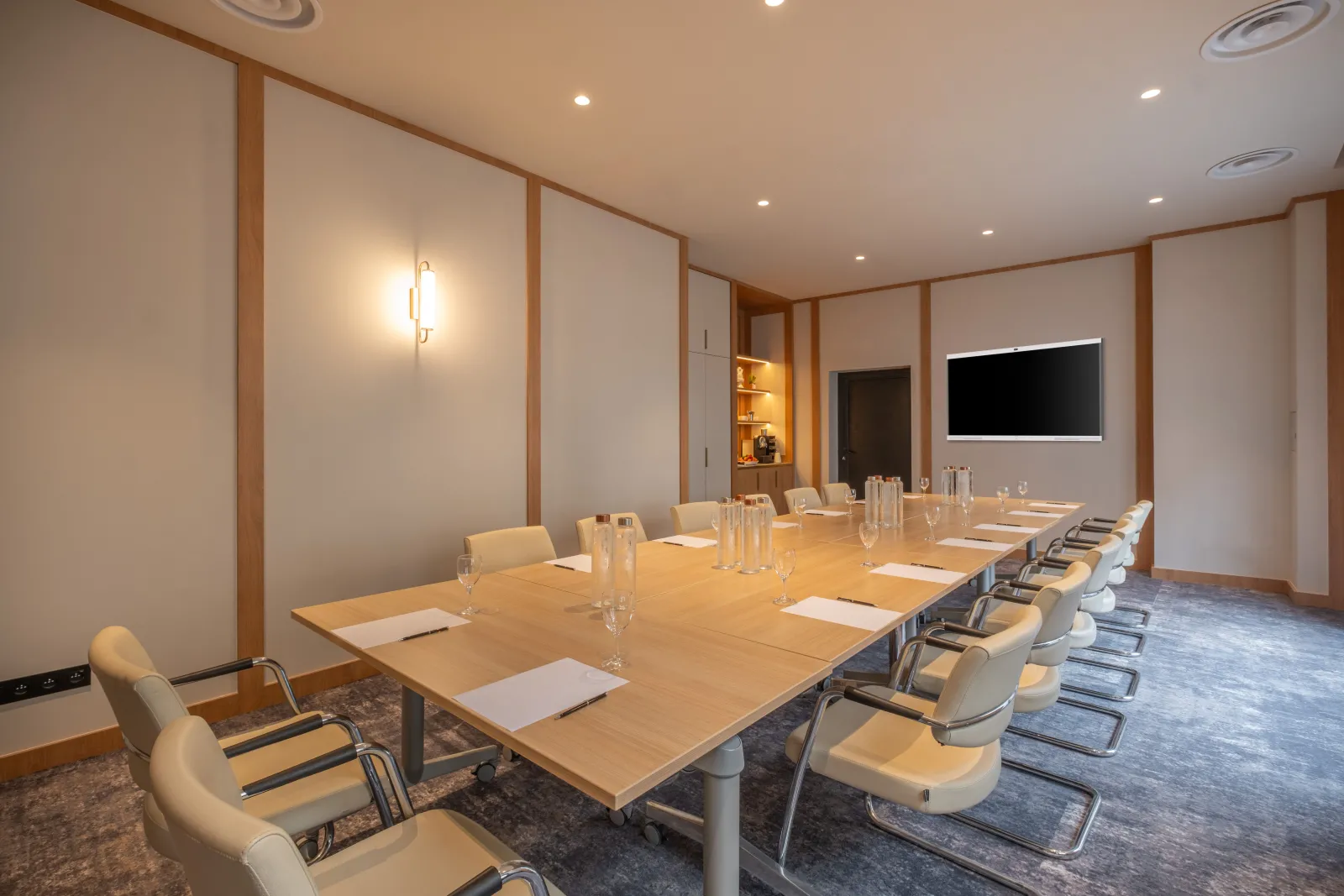 Modern conference room at Burdigala Hotel in Bordeaux with a long wooden table, beige chairs, glass water pitchers, notepads, and a wall-mounted screen, featuring warm lighting and contemporary wood-paneled walls.