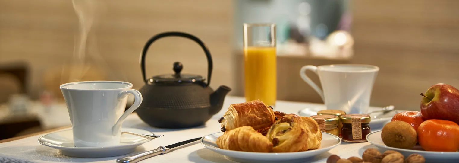 Breakfast table setup with pastries, fresh fruit, orange juice, tea cup, black teapot, and jam jars in a bright room