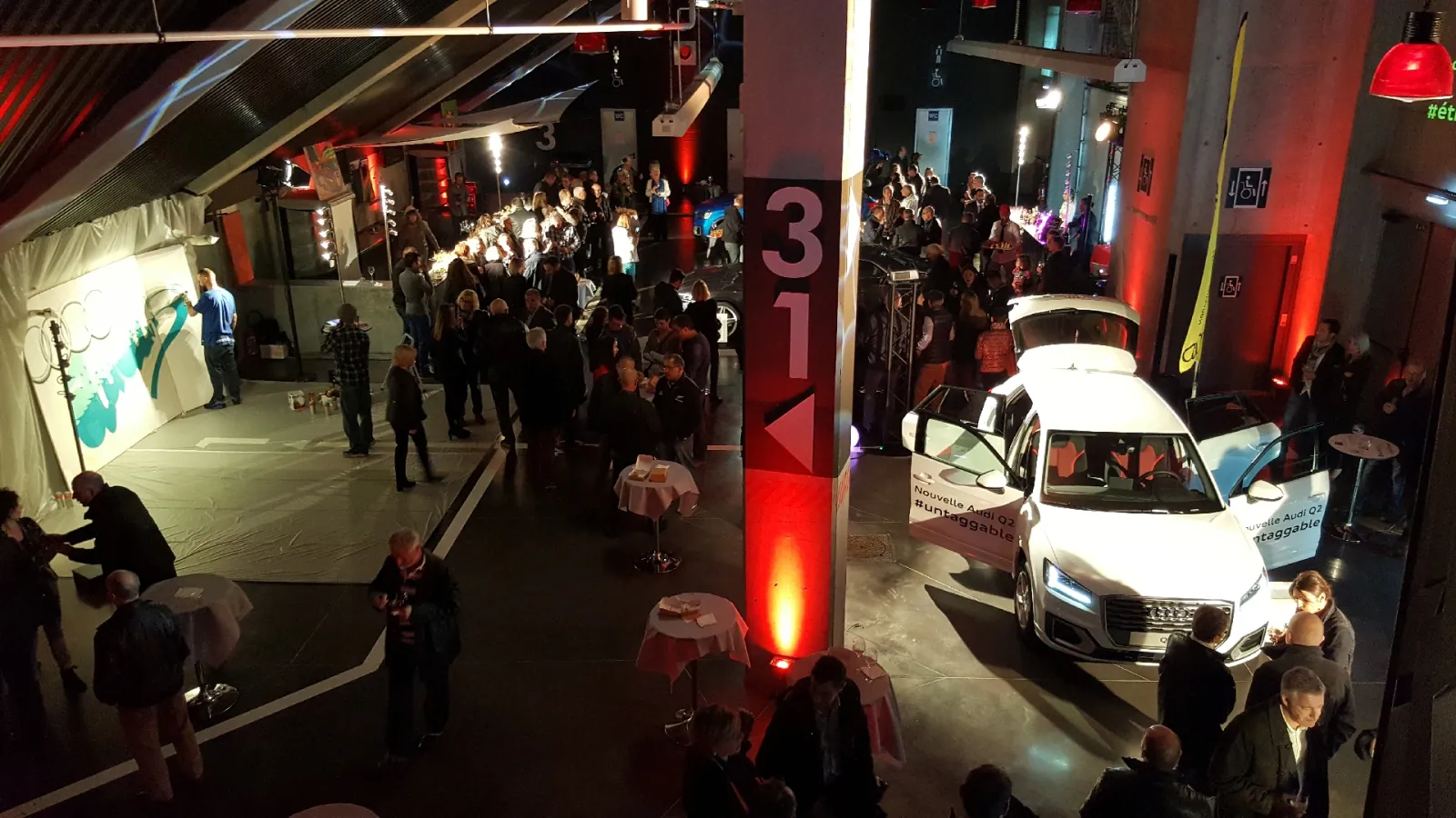 Interior view of a modern event space or exhibition with red ambient lighting, featuring a white car on display, multiple attendees in dark clothing, and interactive installations including a projected screen on the left wall.