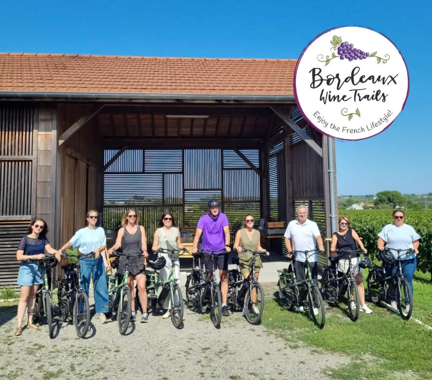 A group of ten cyclists with their bikes standing in front of a modern wooden building overlooking vineyard landscapes in Bordeaux, France.