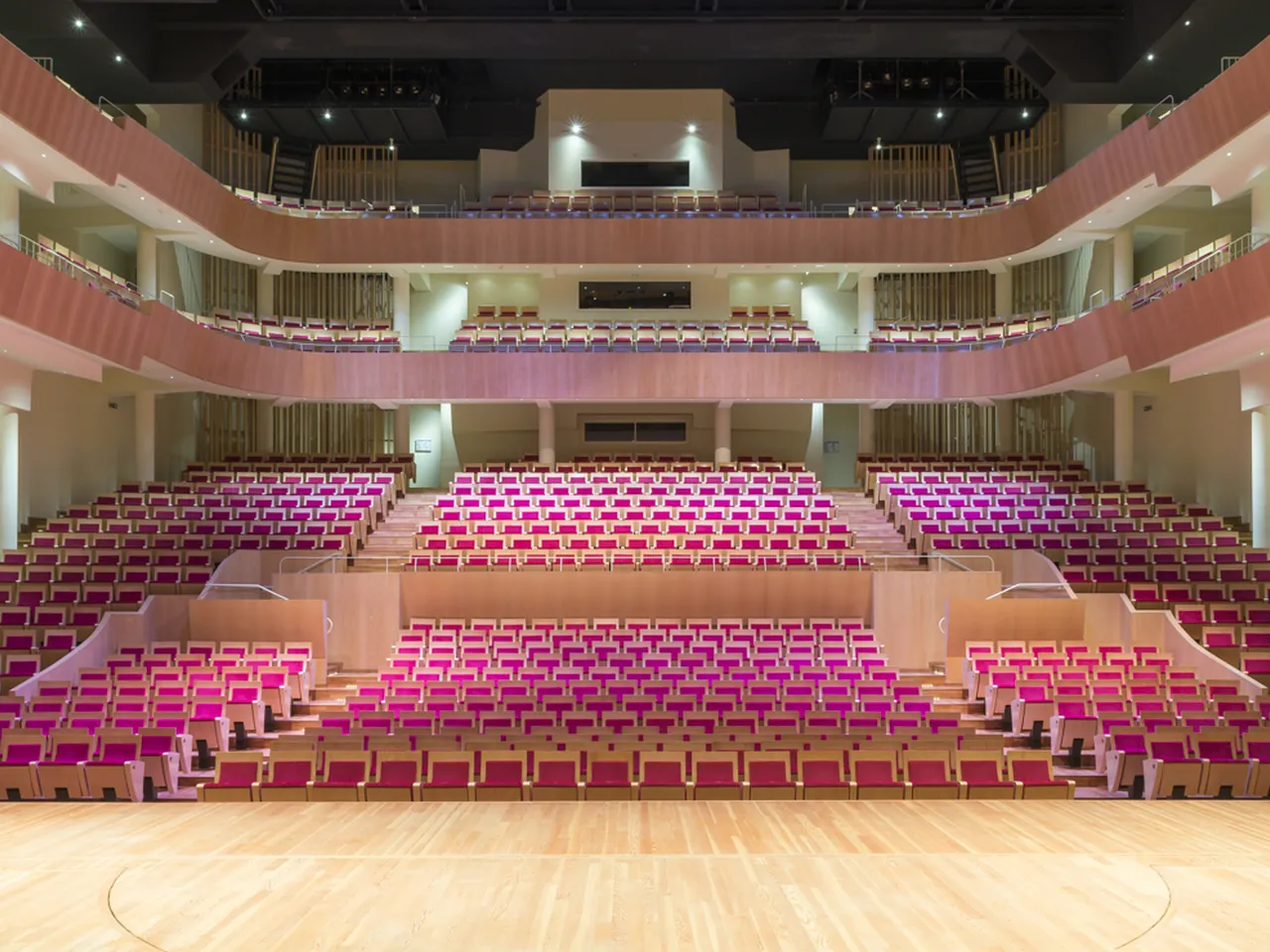 Interior view of the Grand Théâtre of Bordeaux, showing multiple levels of magenta and pink velvet seats arranged in a semicircular amphitheater, with a wooden stage floor in the foreground and ornate architectural details including balconies, railings, a