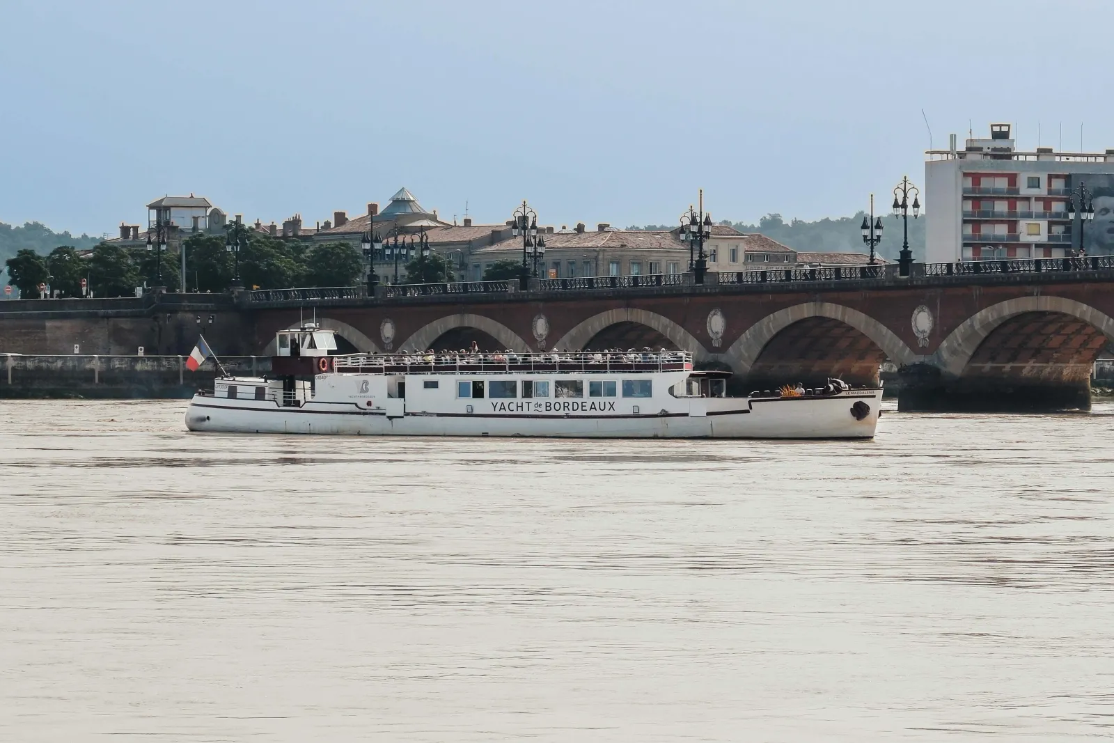 A white river cruise boat named 'Yacht de Bordeaux' sails on the Garonne River in front of the historic Stone Bridge, with 18th-century buildings and architecture visible along the waterfront of Bordeaux, France.