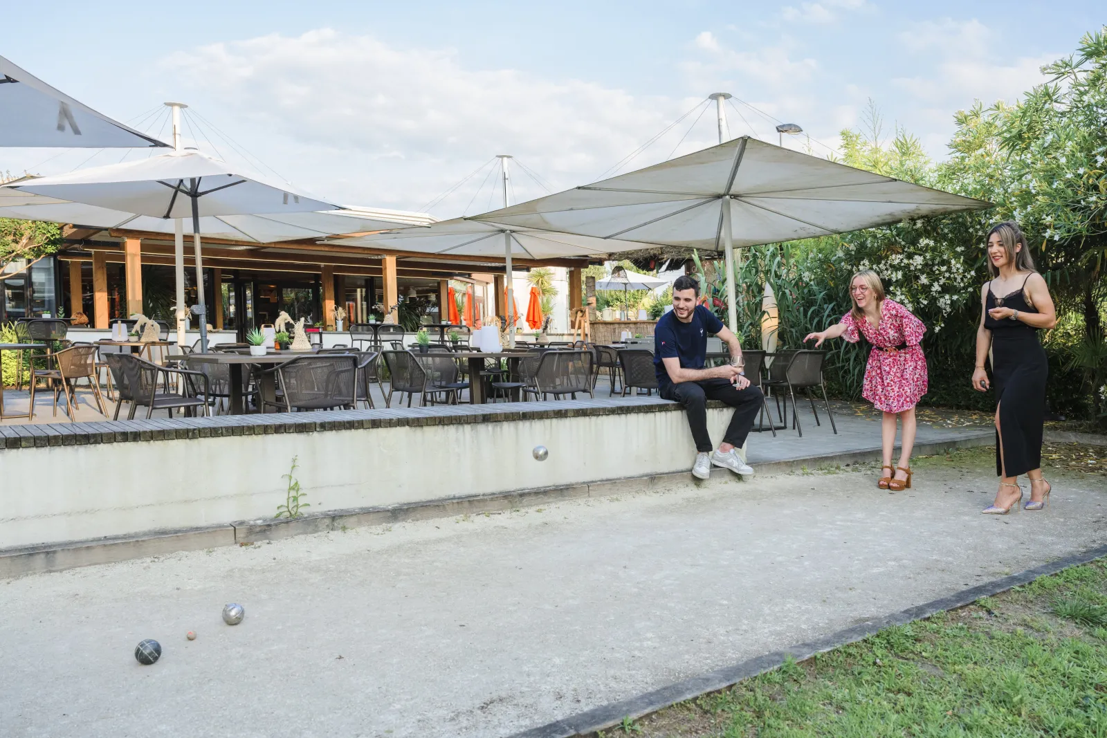 Four people playing boules in the outdoor recreational area of Novotel Bordeaux Lac hotel, with a shaded terrace and restaurant in the background