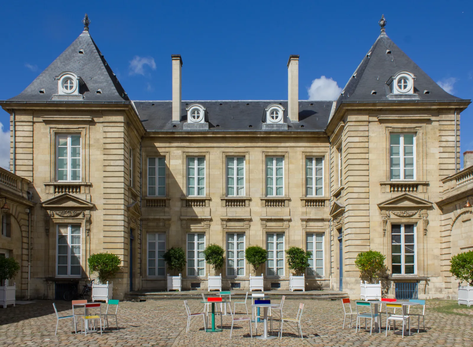 Courtyard of the Musée des Arts Décoratifs et du Design in Bordeaux, featuring a grand 18th-century mansion with mansard roofs, symmetrical architecture, and colorful modern chairs arranged in the stone courtyard on a sunny day.