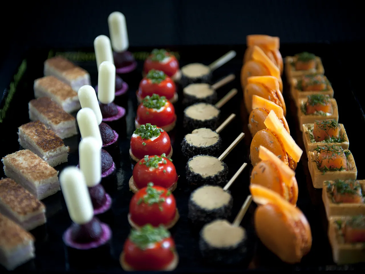 Fine pastry and gourmet appetizers display featuring layered cakes, white cylindrical desserts, red tomato tartlets, black canapés with cream, glazed orange pastries, and herb-topped squares on a black serving board