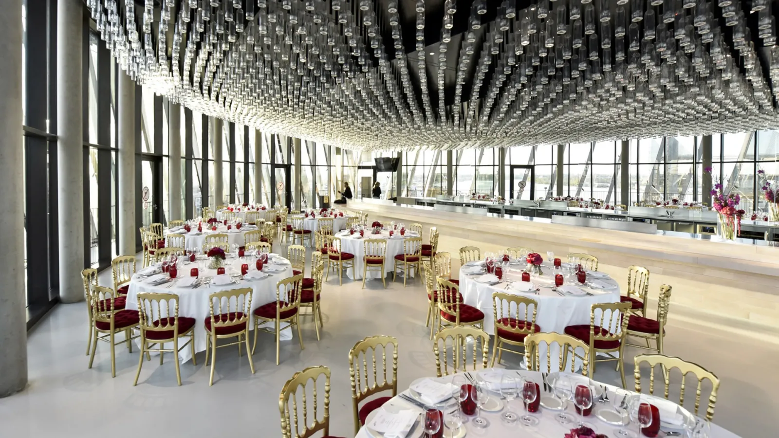 Elegant banquet hall at Cité du Vin in Bordeaux featuring round tables with white linens, gold chairs with burgundy cushions, red floral centerpieces, and a striking geometric ceiling with hanging glass elements, overlooking the city.