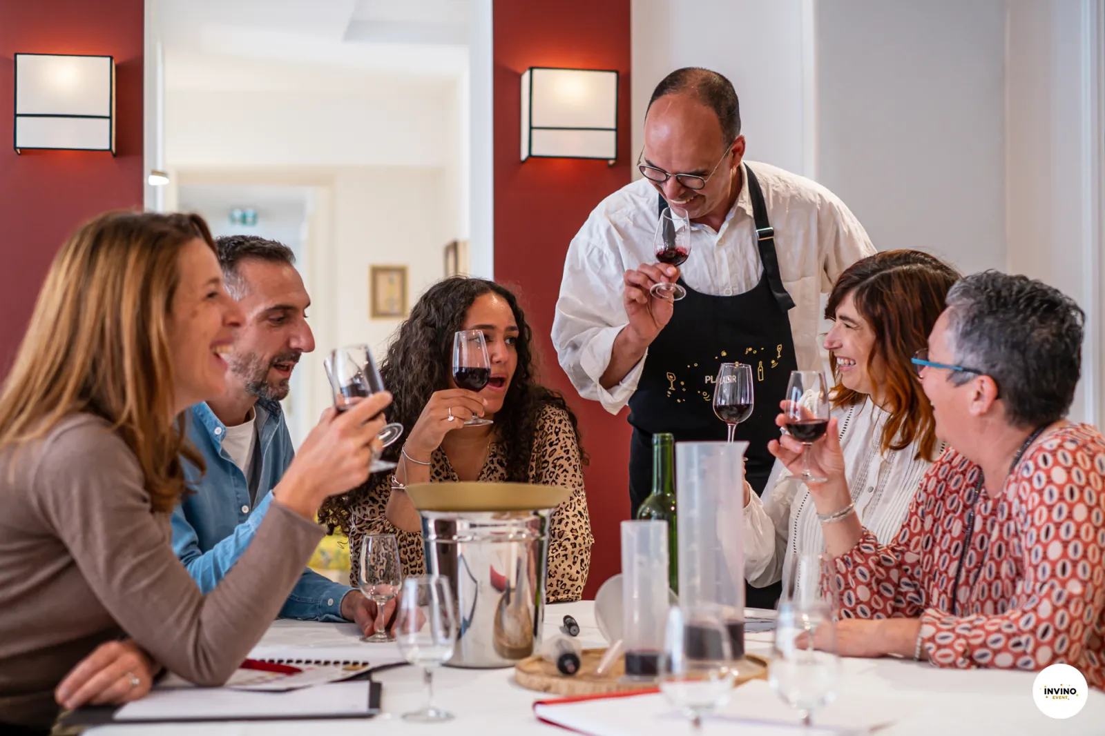 A sommelier in a black apron presents wine to a group of five adults seated at a table during a wine tasting event in Bordeaux, with red and white walls and wine glasses on the table.