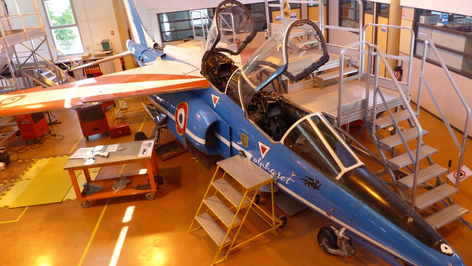 Blue and white jet-powered racing aircraft displayed in an indoor hangar at Aérocampus Aquitaine in Bordeaux, with open cockpit canopies and maintenance stairs visible