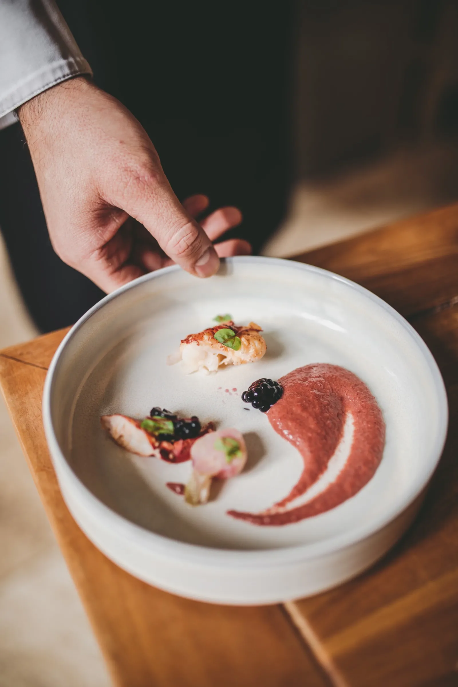 A chef in white shirt presenting an elegantly plated fine dining dish on a white plate with artistic food arrangement including seafood, garnishes, and red sauce swirl on a wooden table.
