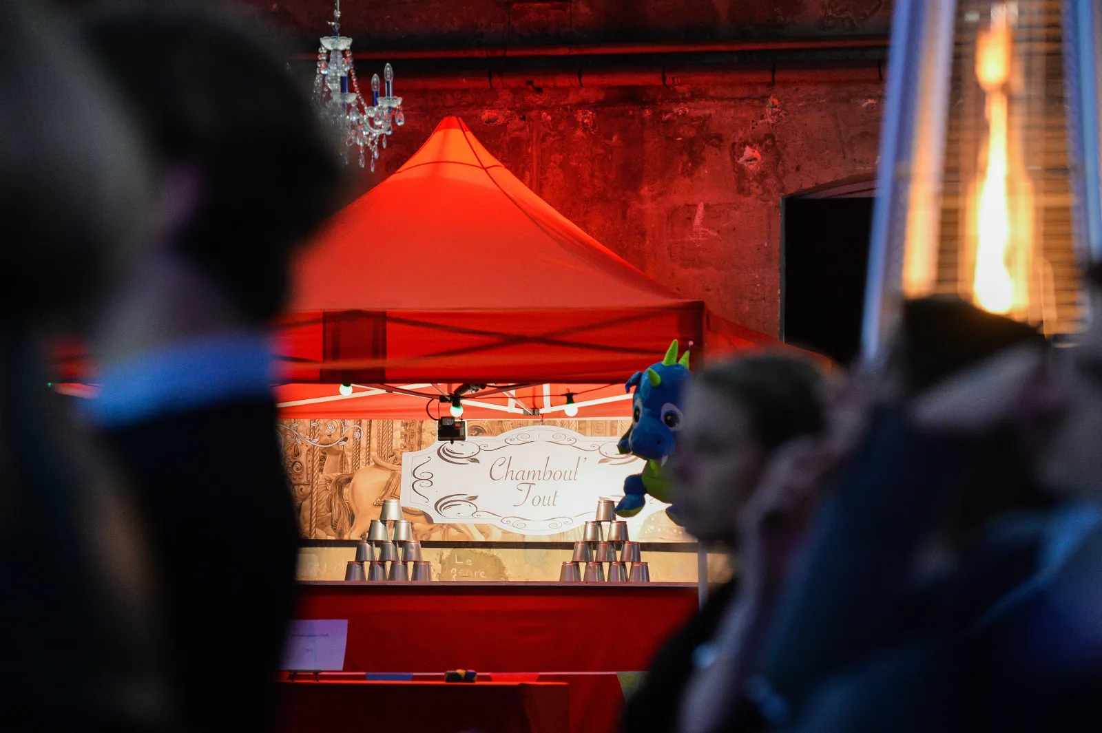 A festive market stall with a red tent canopy displaying 'Chamboul Tout' sign, decorated with a colorful peacock figure, surrounded by stacked cups and visitors in Bordeaux's Christmas market