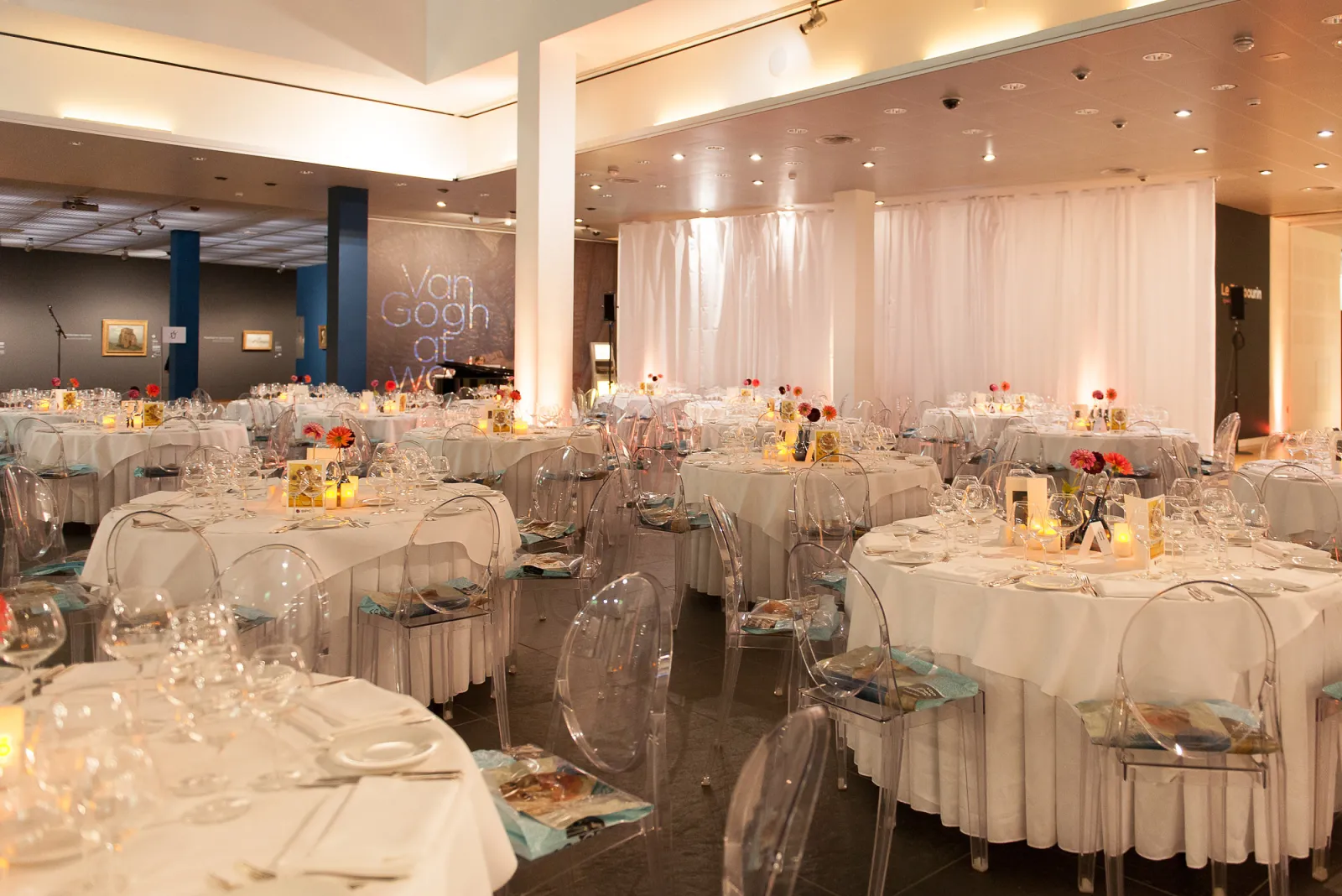 Elegant banquet hall in Bordeaux with round tables draped in white tablecloths, transparent chairs, floral centerpieces, and white curtains, set for a formal event at Cézame Atlantique.