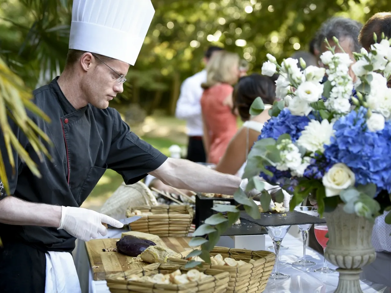 A chef in white hat and black chef's coat prepares tamales at an outdoor catering event in Bordeaux, with white and blue floral arrangements and guests visible in the background.