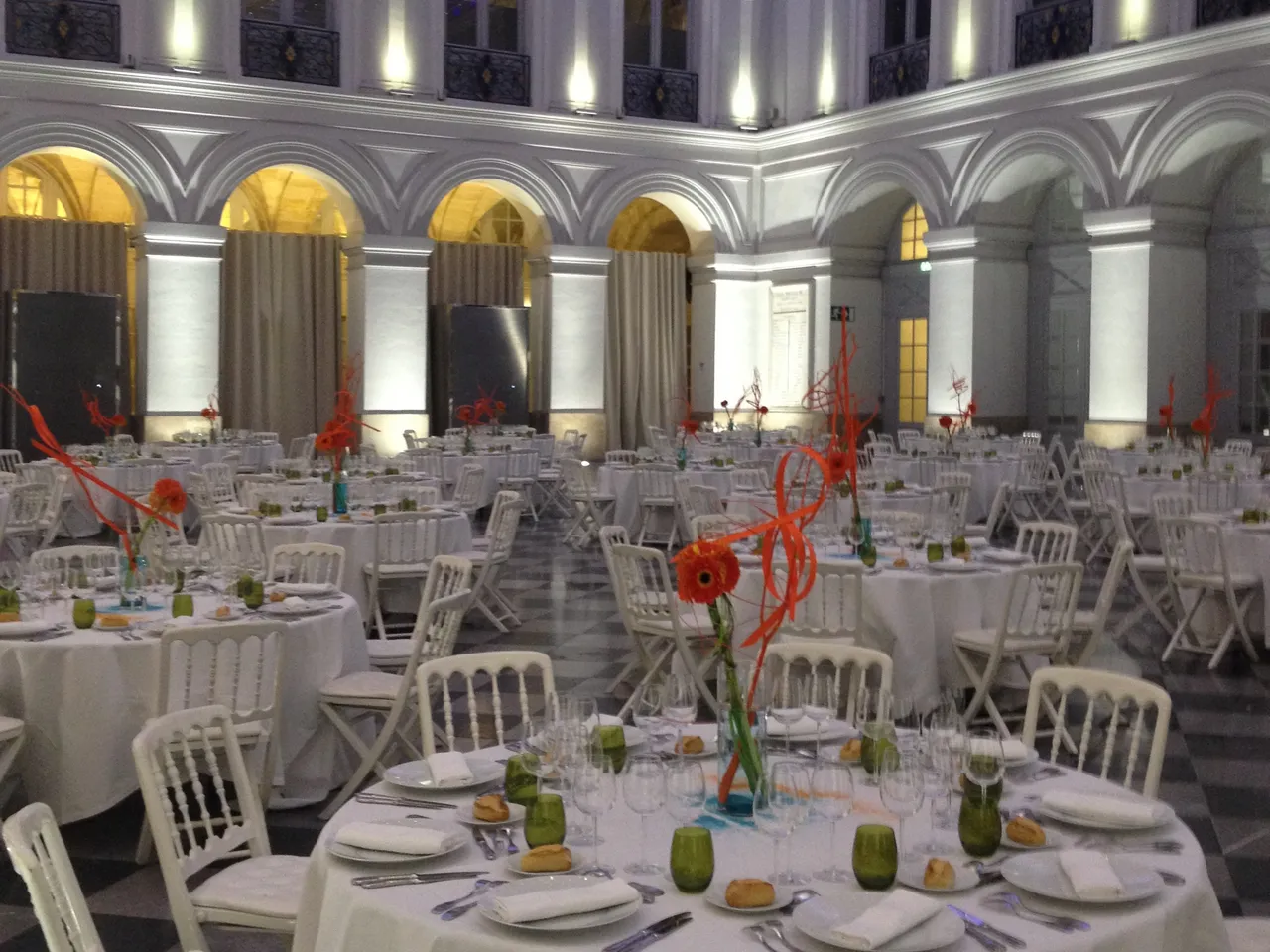 Elegant banquet hall with white round tables, white chairs, and red floral centerpieces in a classical architecture setting with arched columns and warm lighting