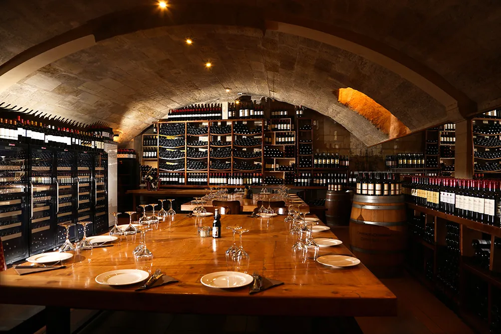 Underground wine cellar with arched stone ceiling, long wooden dining table set for service, wine glasses and place settings, extensive wine bottle storage shelves lining the walls, warm ambient lighting.