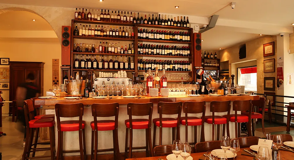 Interior of a Bordeaux brasserie featuring a wooden bar with red cushioned stools, extensive wine bottle display on illuminated shelves behind the counter, and elegant dining area with set tables in warm ambient lighting.