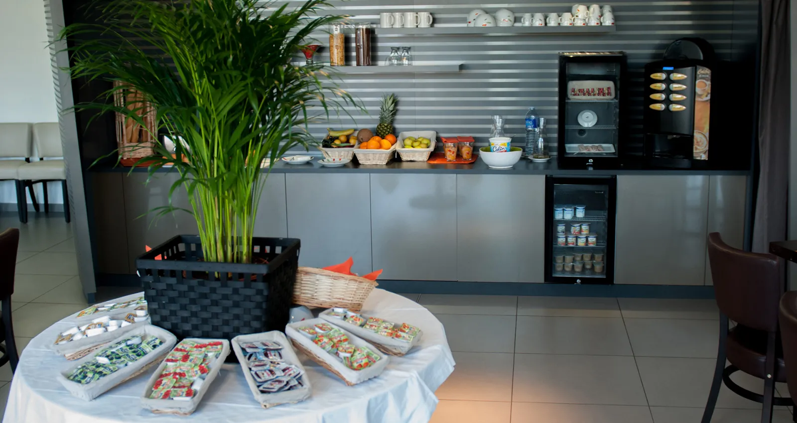 Modern breakfast area in an apartment hotel featuring a white table with pastries and treats, a potted plant, and a contemporary gray kitchenette with coffee machine, refrigerator, and shelving with dishware and snacks in the background.