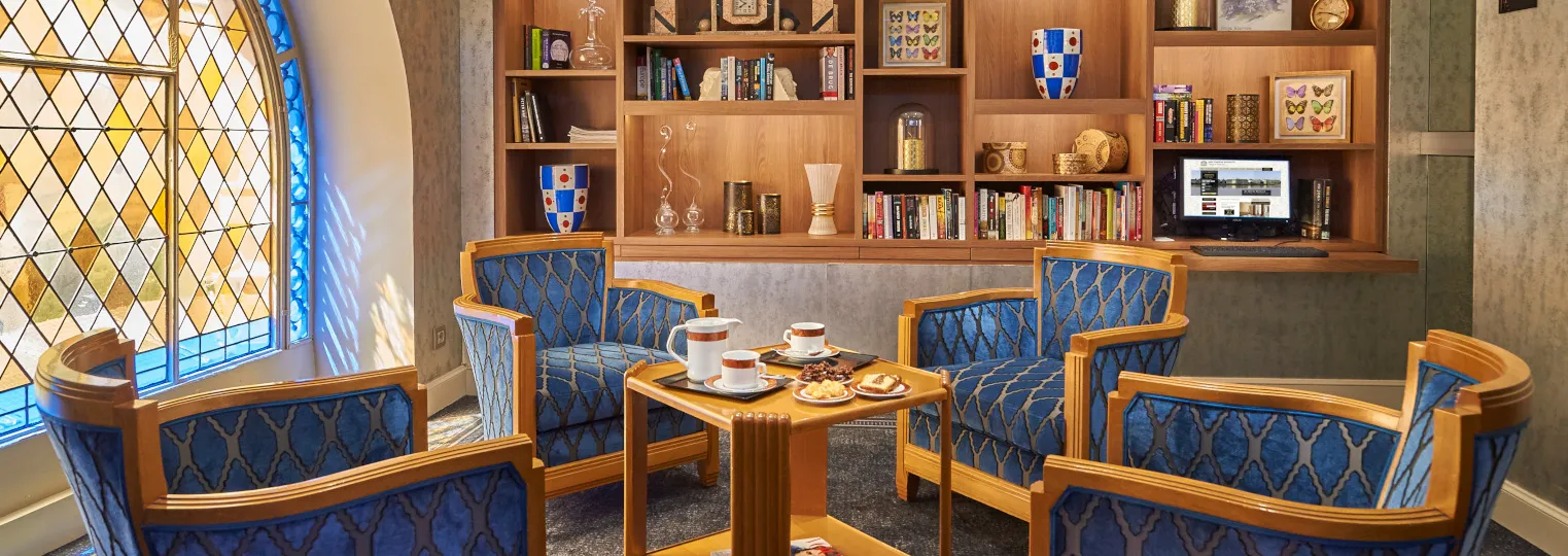 Elegant hotel lounge at Best Western Premier Hôtel Bayonne Etche Ona in Bordeaux, featuring blue upholstered wooden armchairs, a small coffee table with refreshments, extensive wooden bookshelves, a leaded glass window, and decorative elements including b