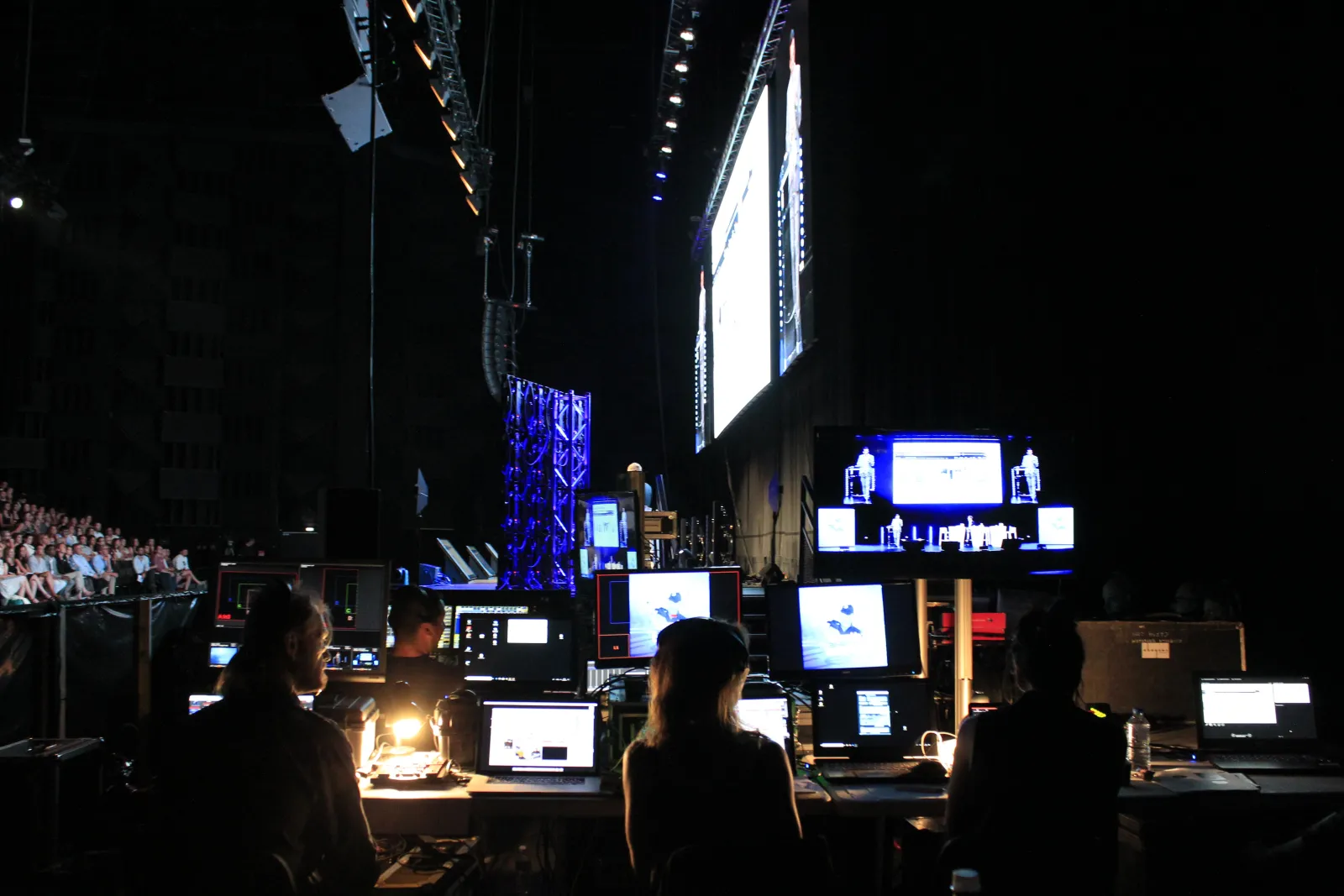 Technical crew operating lighting and projection equipment at a large indoor event venue in Bordeaux, with multiple screens displaying content and an audience visible in the background.