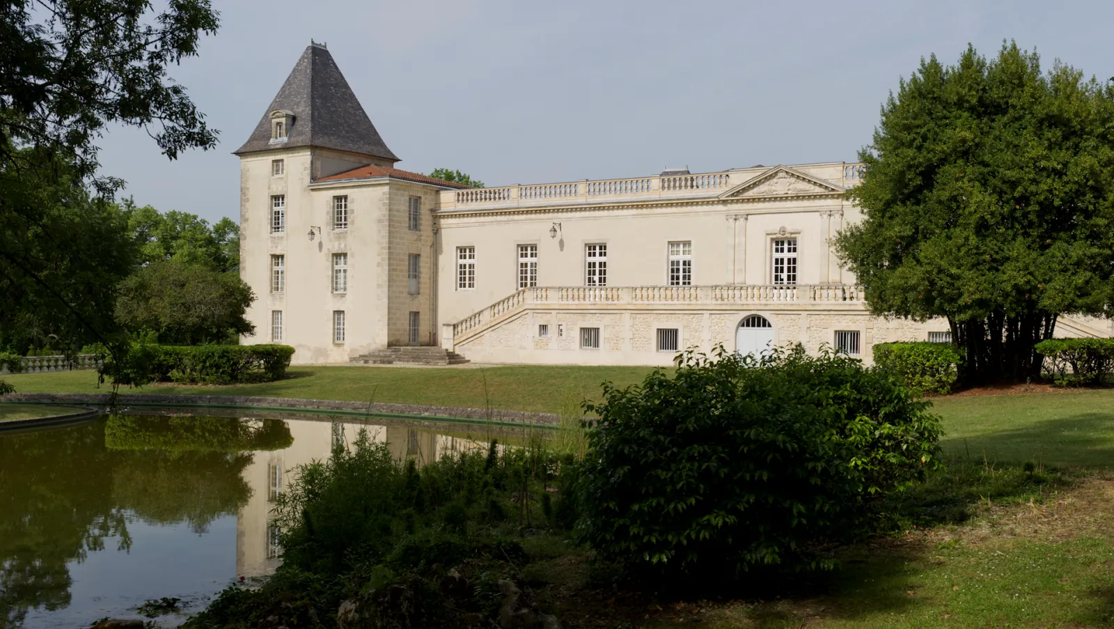 A white neoclassical château with a conical tower, surrounded by manicured lawns and trees, with a reflecting pond in the foreground.