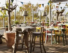 Outdoor terrace of Mama Shelter Bordeaux with wooden tables and chairs arranged among trees, featuring string lights and a relaxed atmosphere in a public garden setting.