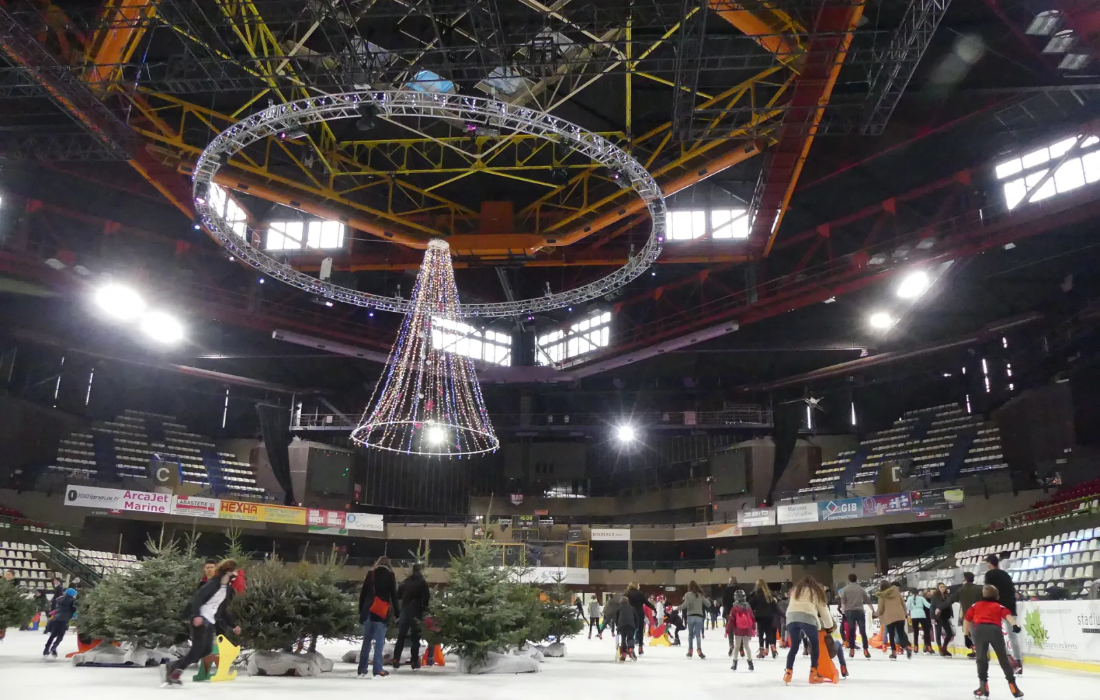 Indoor ice skating rink at Mériadeck arena in Bordeaux with visitors skating, decorated with Christmas lights and garland, under industrial ceiling with yellow metal beams and orange cranes