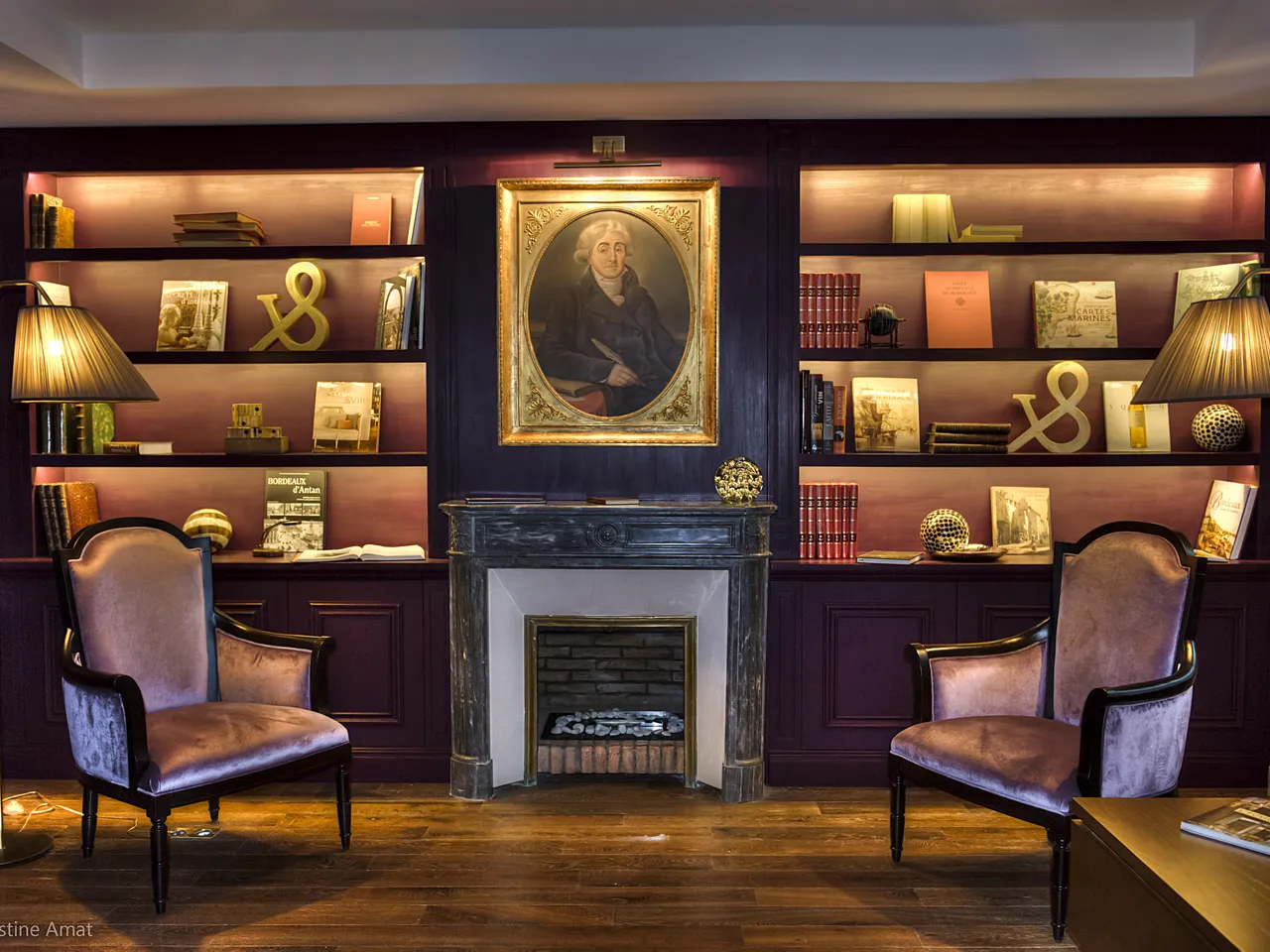 Elegant library interior at Hôtel de Sèze in Bordeaux, featuring dark wood paneling with built-in bookshelves, a classical portrait above a fireplace mantel, two burgundy leather armchairs, brass decorative elements, and warm ambient lighting.