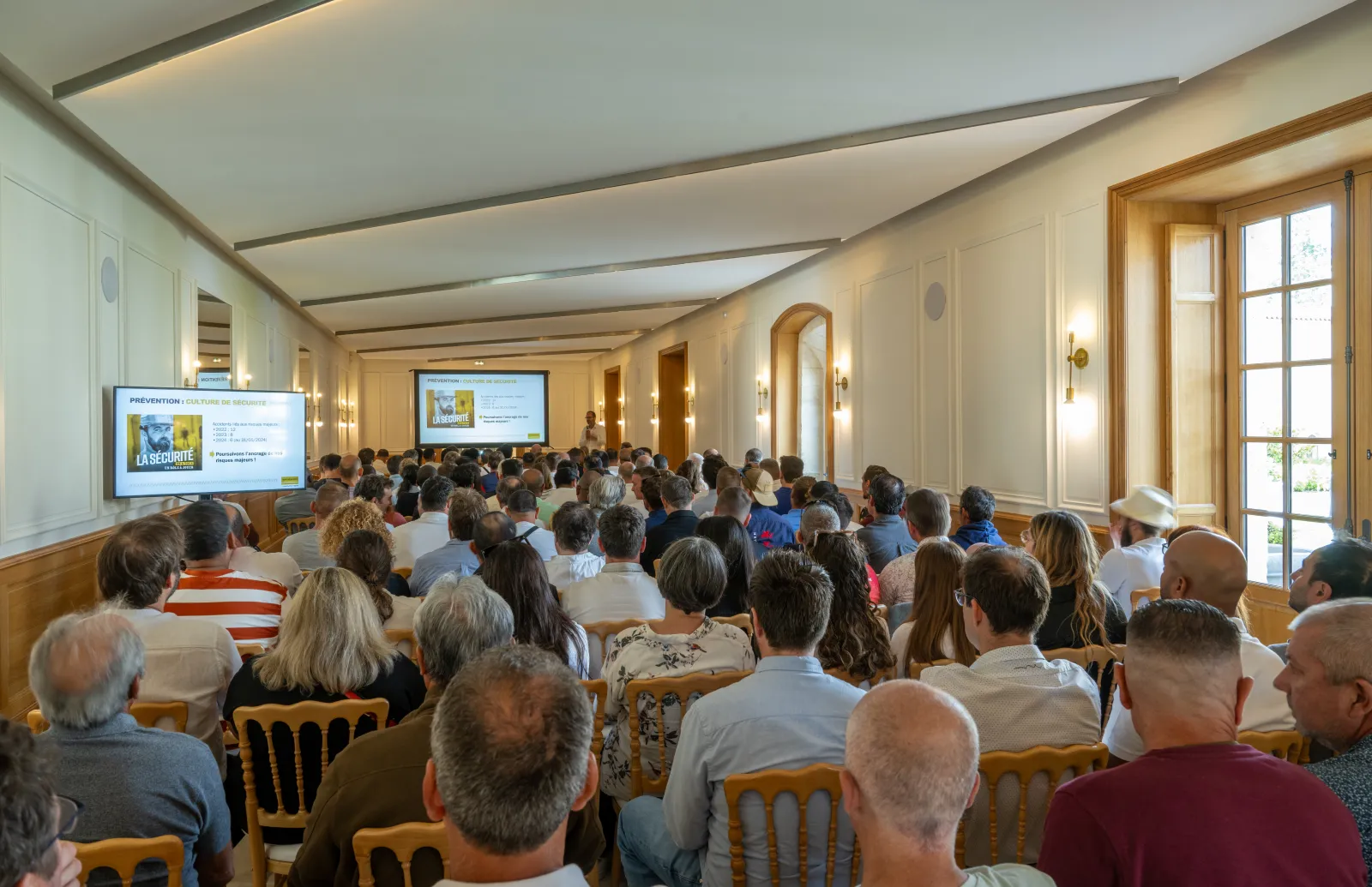 Large audience seated in rows attending a presentation in an elegant conference hall with wooden chairs, cream-colored walls, arched doorways, and projection screens displaying content about La Segonde.