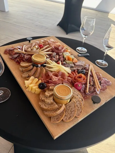 Elegant charcuterie and appetizer board on wooden platter with cured meats, cheeses, bread, spreads, and garnishes, served at a rooftop venue with wine glasses on a black table