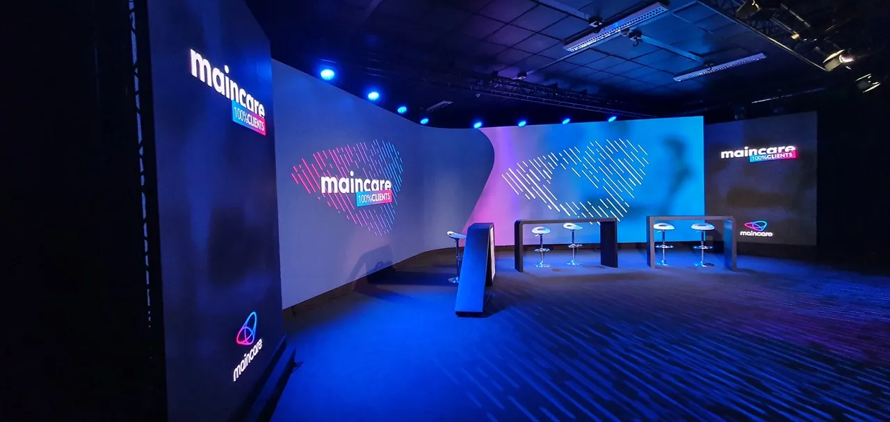 Modern event space with blue and purple lighting, featuring Maincare branding on large displays and conference seating areas