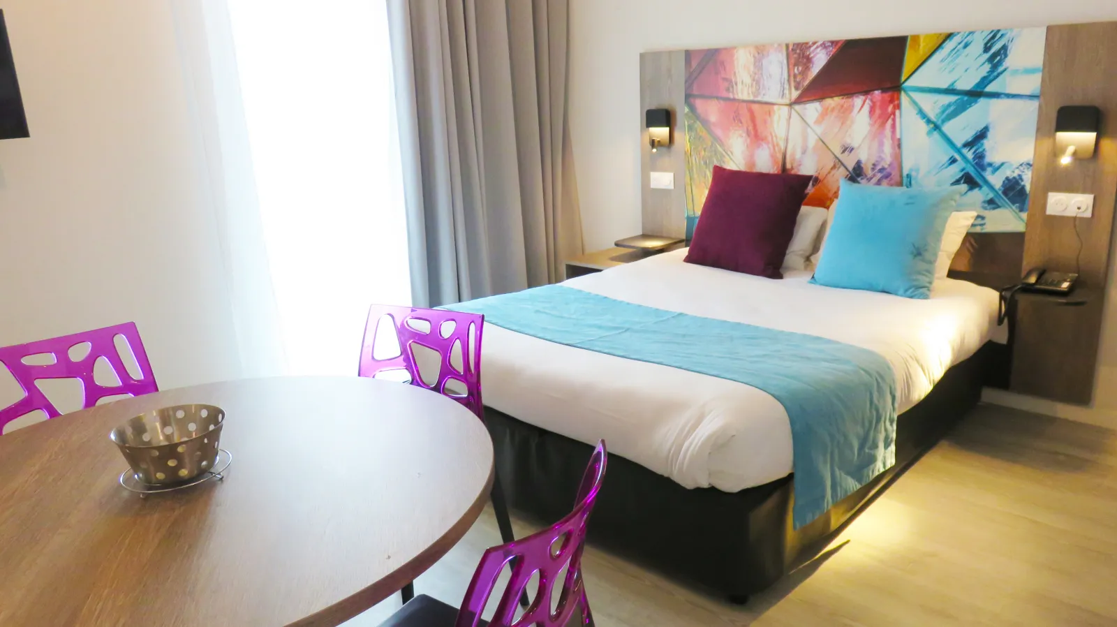Modern hotel room with a queen bed, colorful abstract artwork headboard, purple designer chairs at a round table, floor-to-ceiling windows with curtains, and contemporary decor in blue and warm tones.