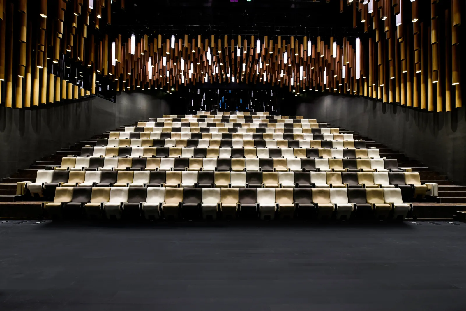 Interior view of a modern cinema auditorium with rows of beige and black seats arranged in ascending tiers, illuminated by warm golden lighting and contemporary ceiling fixtures.