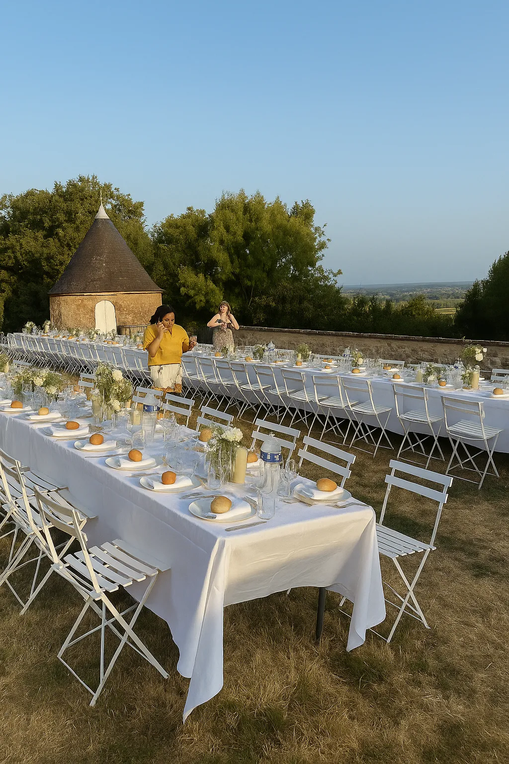 Outdoor wedding reception setup on a terrace with long white-draped tables, white chairs, and orange place card holders, overlooking a countryside landscape with a historic stone tower and trees.