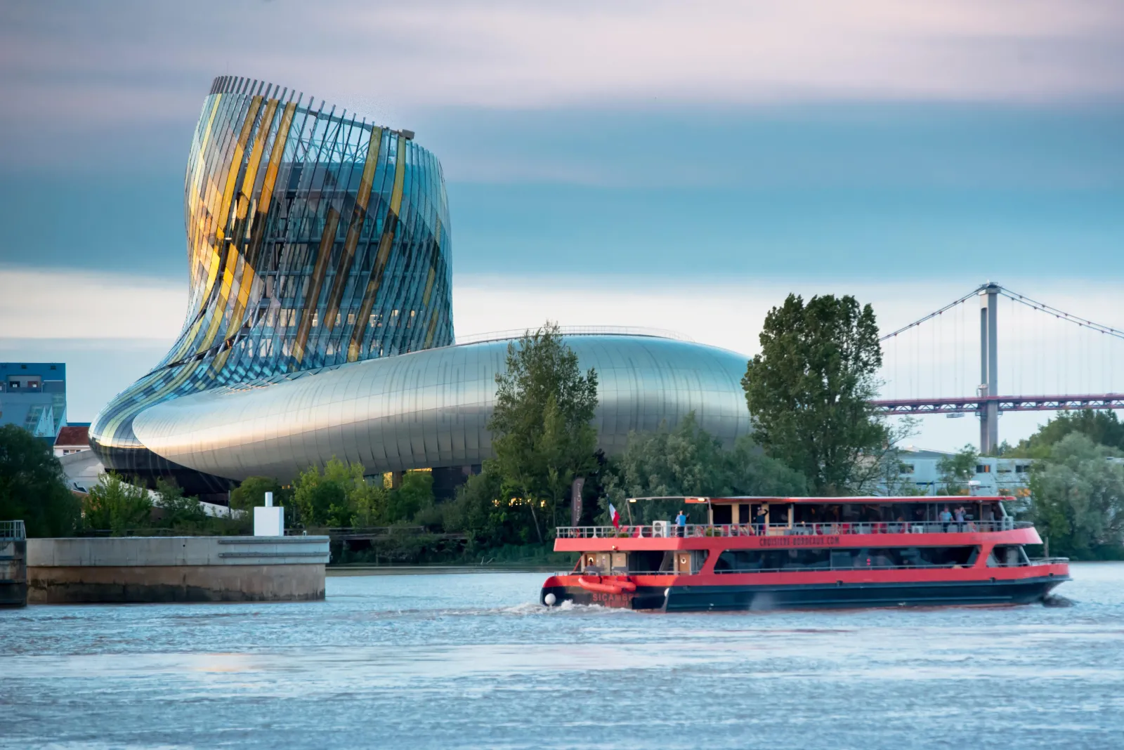 A red passenger riverboat navigates the Garonne River in front of the modern curved architecture of the Cité du Vin wine museum, with the Aquitaine Bridge visible in the background under a blue sky.