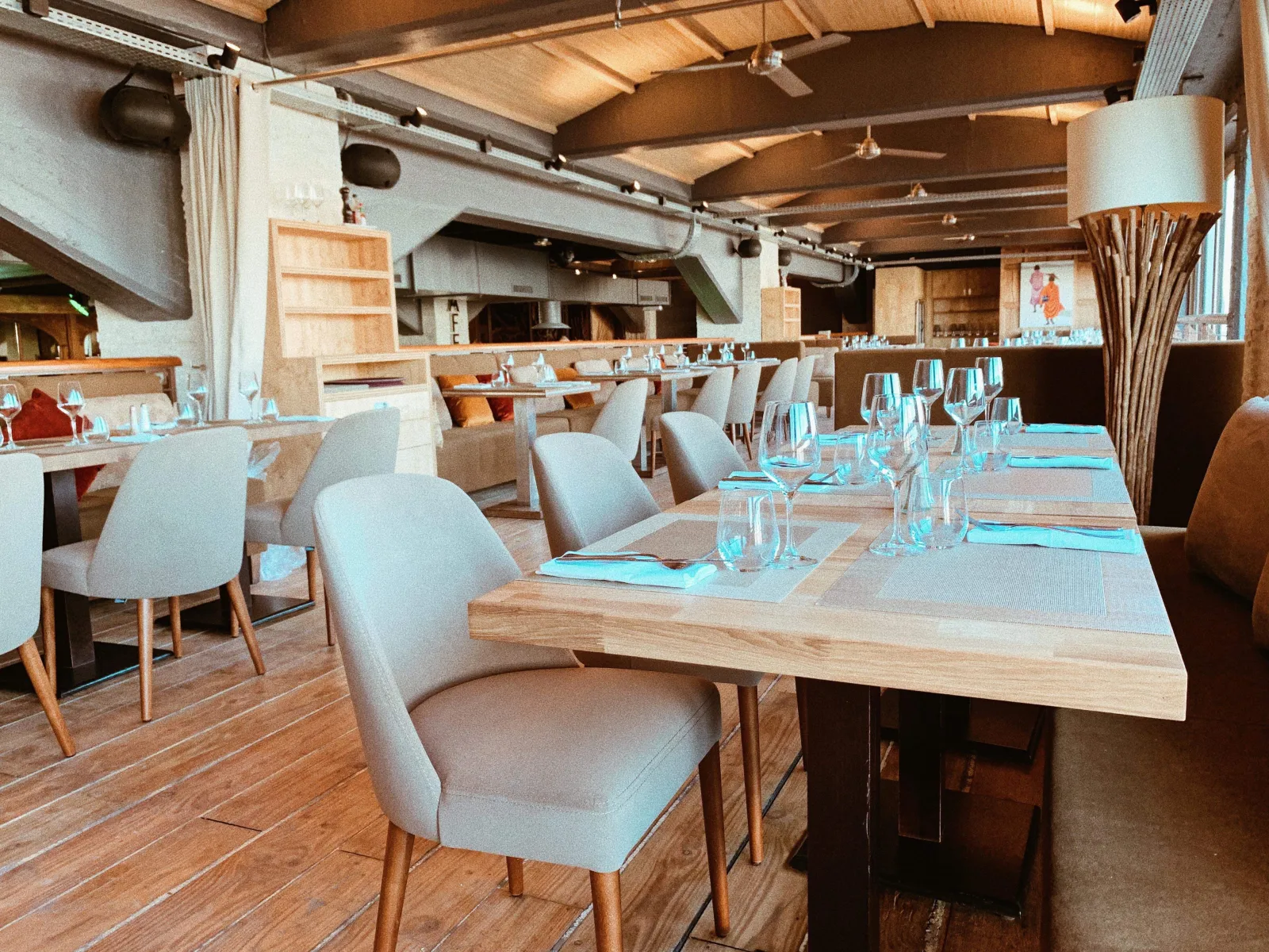 Modern restaurant interior with wooden beamed ceiling, light gray upholstered chairs, natural wood tables set with glassware and blue napkins, and an open kitchen visible in the background at Café Maritime in Bordeaux.