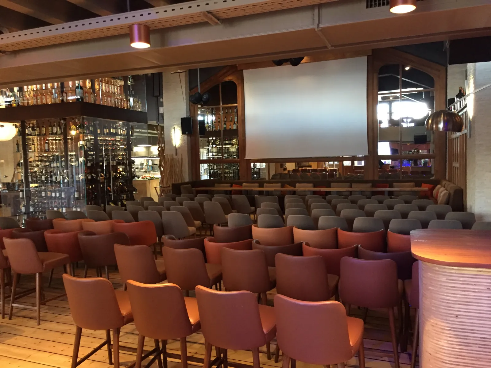 Interior of Café Maritime in Bordeaux featuring rows of coral and gray upholstered chairs facing a projection screen, with a well-stocked bar, wooden beams, and modern lighting in a spacious event venue.