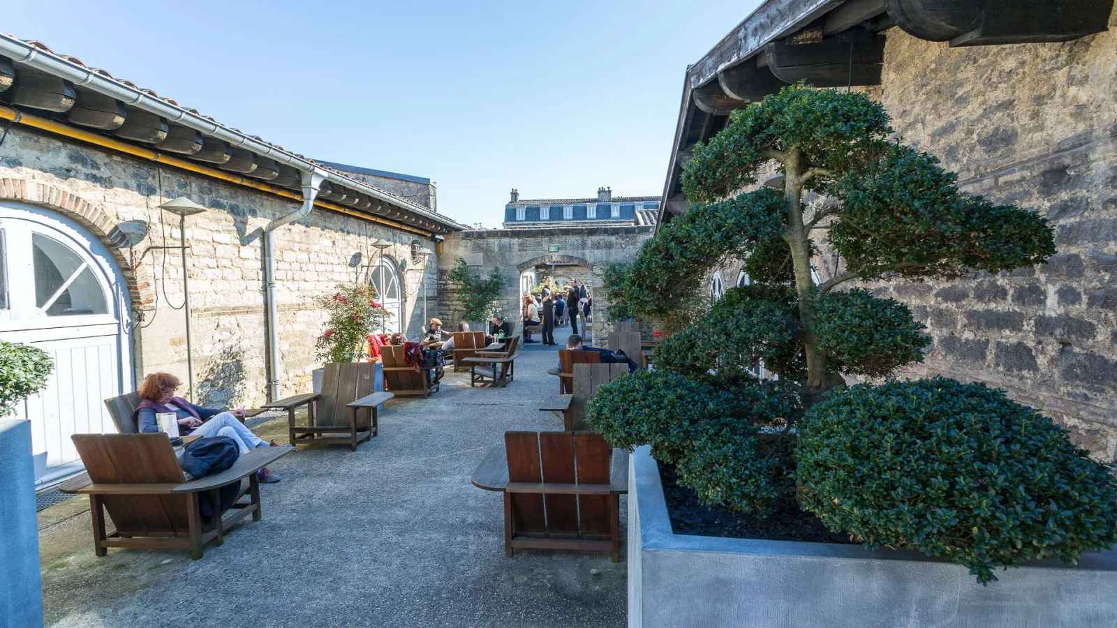 Outdoor courtyard of the CAPC Museum in Bordeaux featuring stone buildings, wooden armchairs, ivy-covered walls, and visitors relaxing in a contemporary art space.