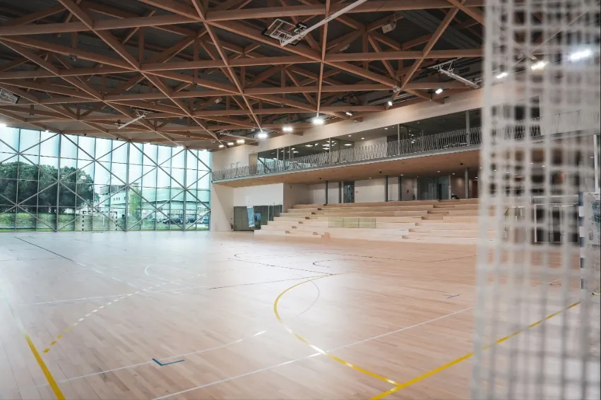 Modern sports hall interior at the University of Bordeaux with wooden flooring, marked court lines, exposed steel roof structure, large windows, and tiered seating area
