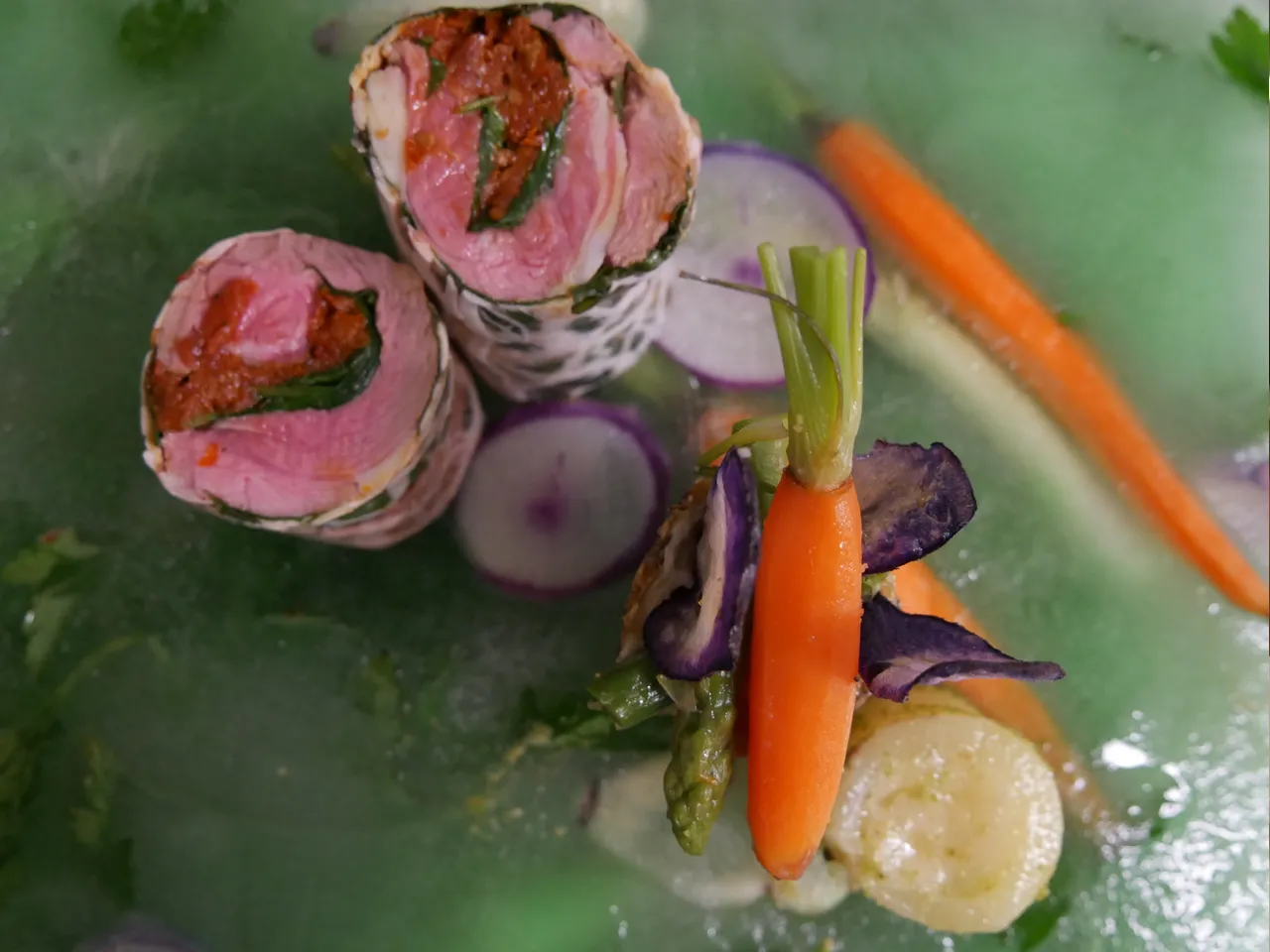 Gourmet appetizer platter featuring sliced beef carpaccio rolls with herbs, purple flowers, orange carrot sticks, and cream sauce on green plate