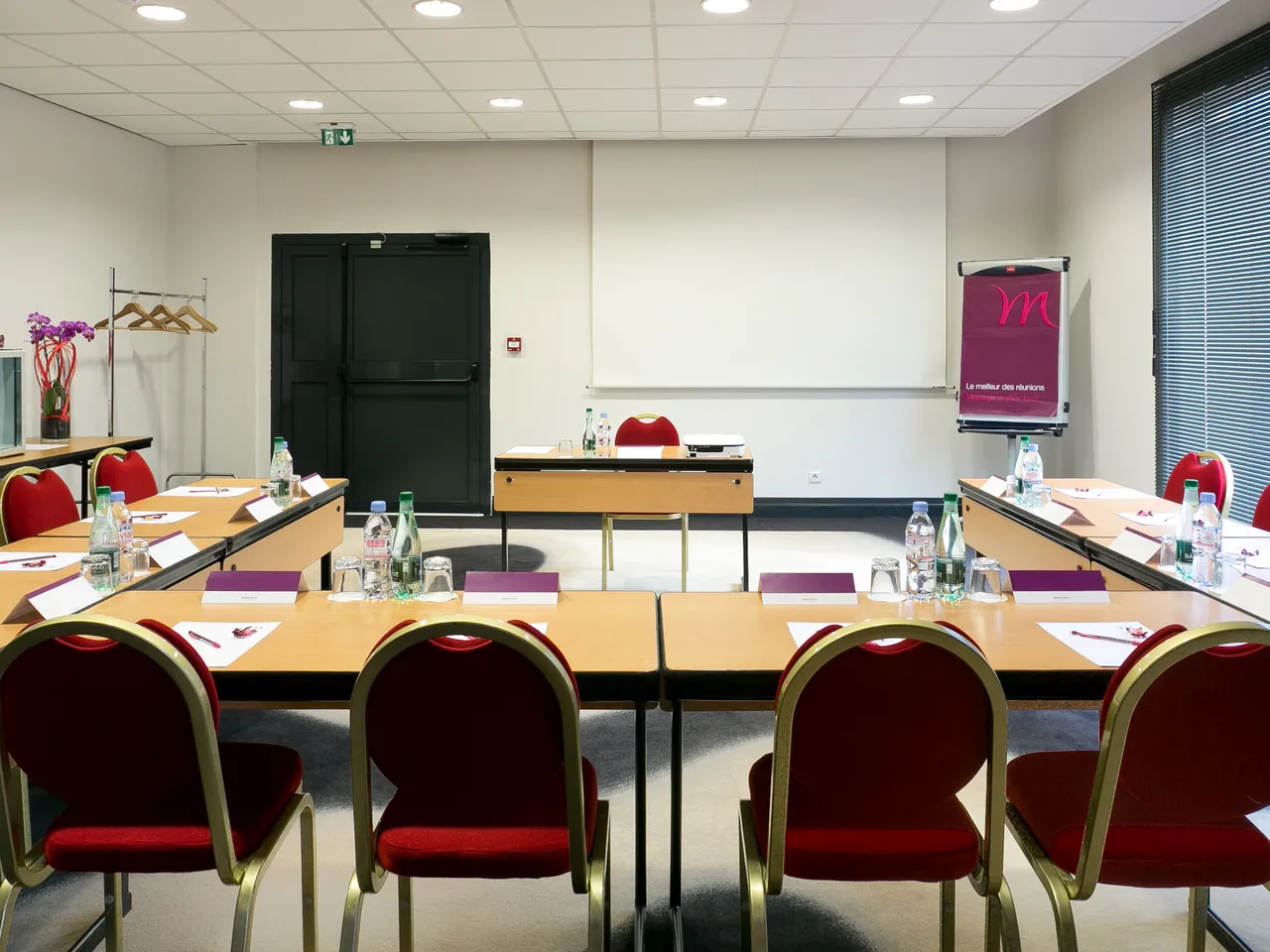 Modern conference room with U-shaped table setup, red chairs, wooden tables, whiteboard, water bottles, and meeting materials at Mercure Bordeaux-Lac hotel.