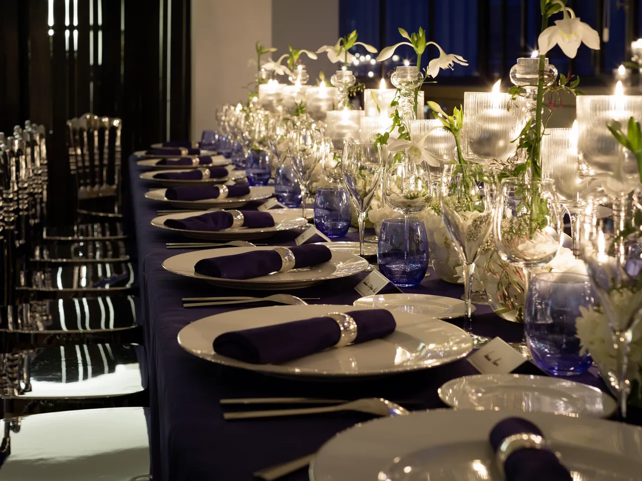 Elegant formal dinner table setting with white plates, dark navy napkins, blue glassware, clear glass vases with white flowers, and candles along a navy tablecloth in a modern event space.